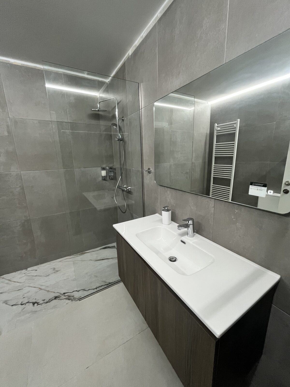 A bathroom in grey stone tile, with a walk-in rain shower and a backlit mirror over the vanity.