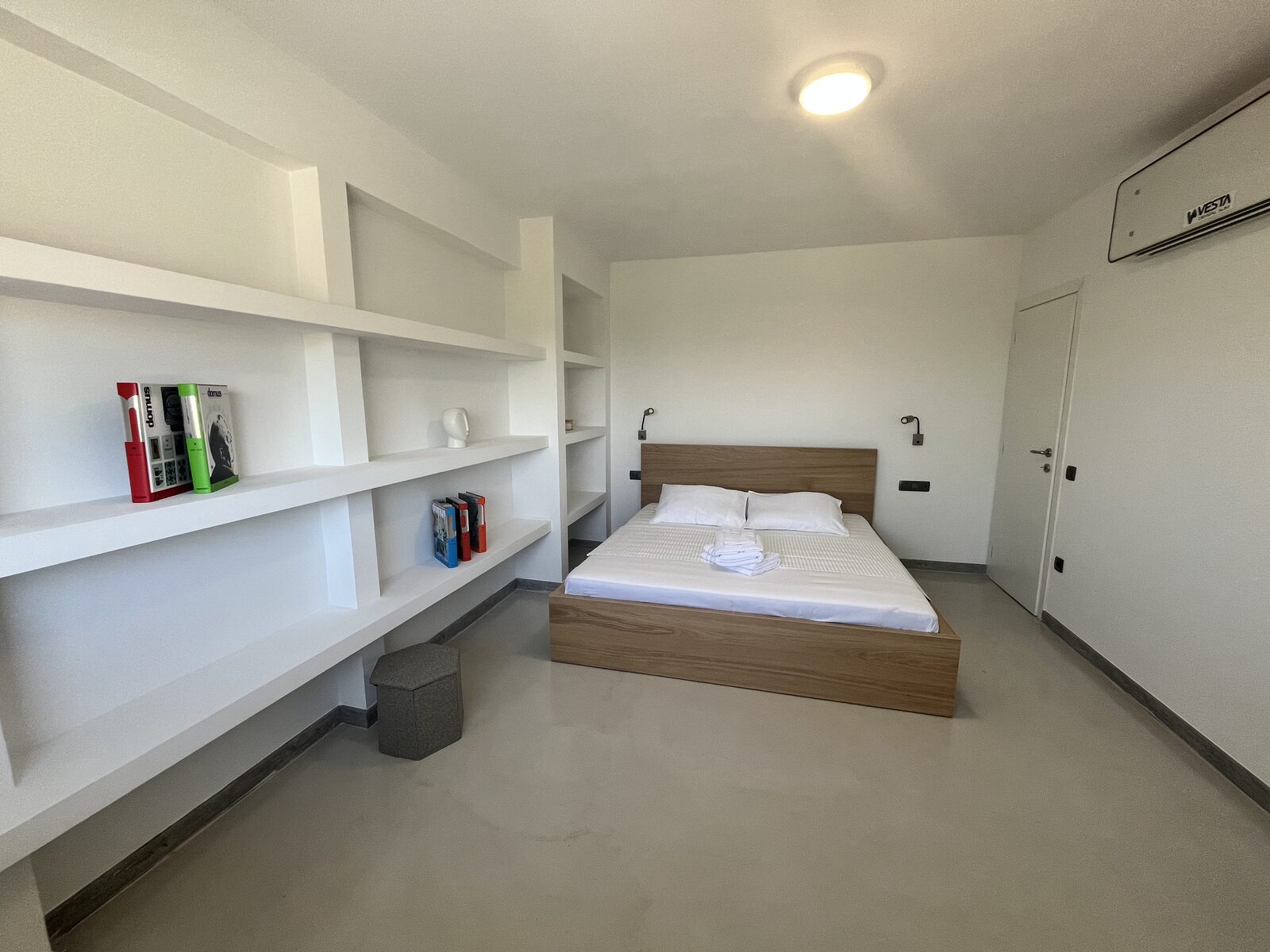 A double bedroom lined with long white shelving, the bed made up in white against pale timber.