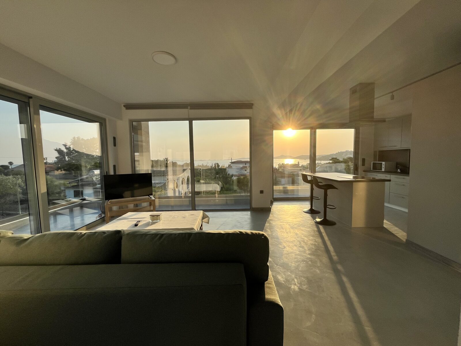 The upper apartment's open-plan living room and kitchen bar at sunset, the sun dropping into the gulf beyond the glass.