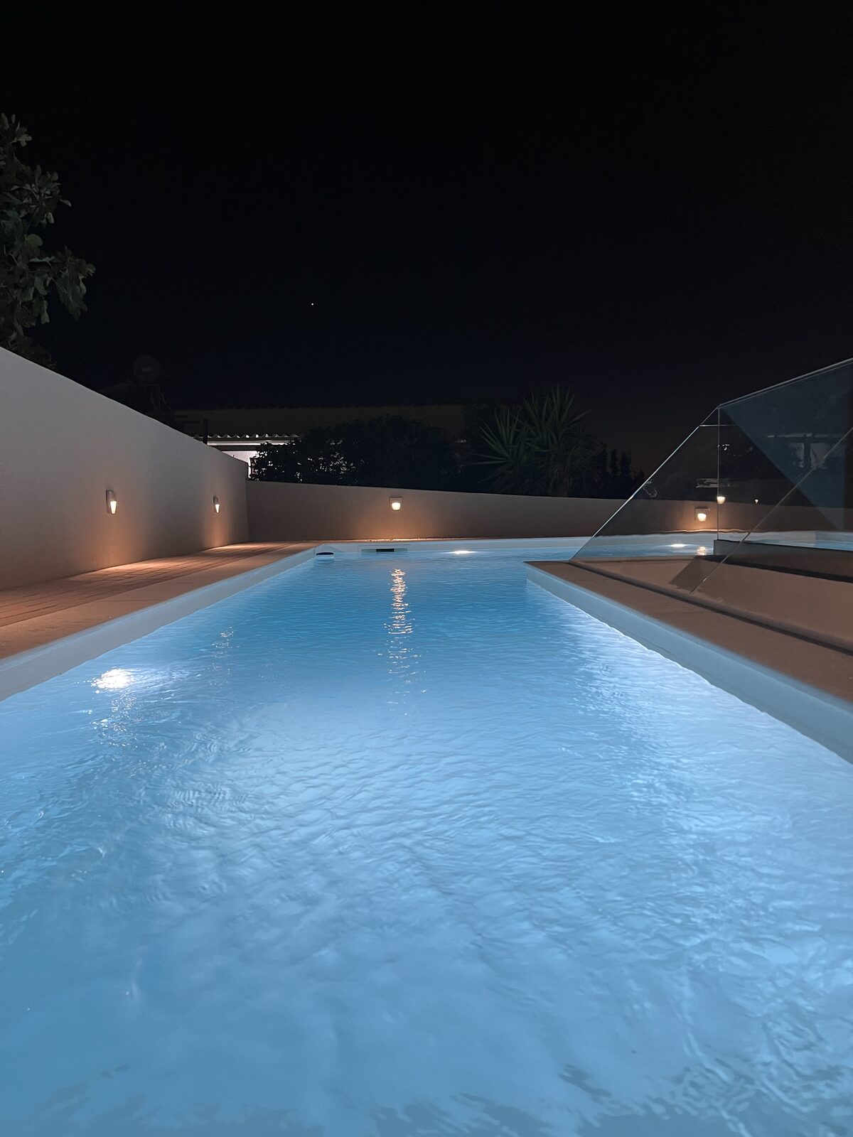 The pool after dark, water glowing pale blue between the deck and the garden wall.